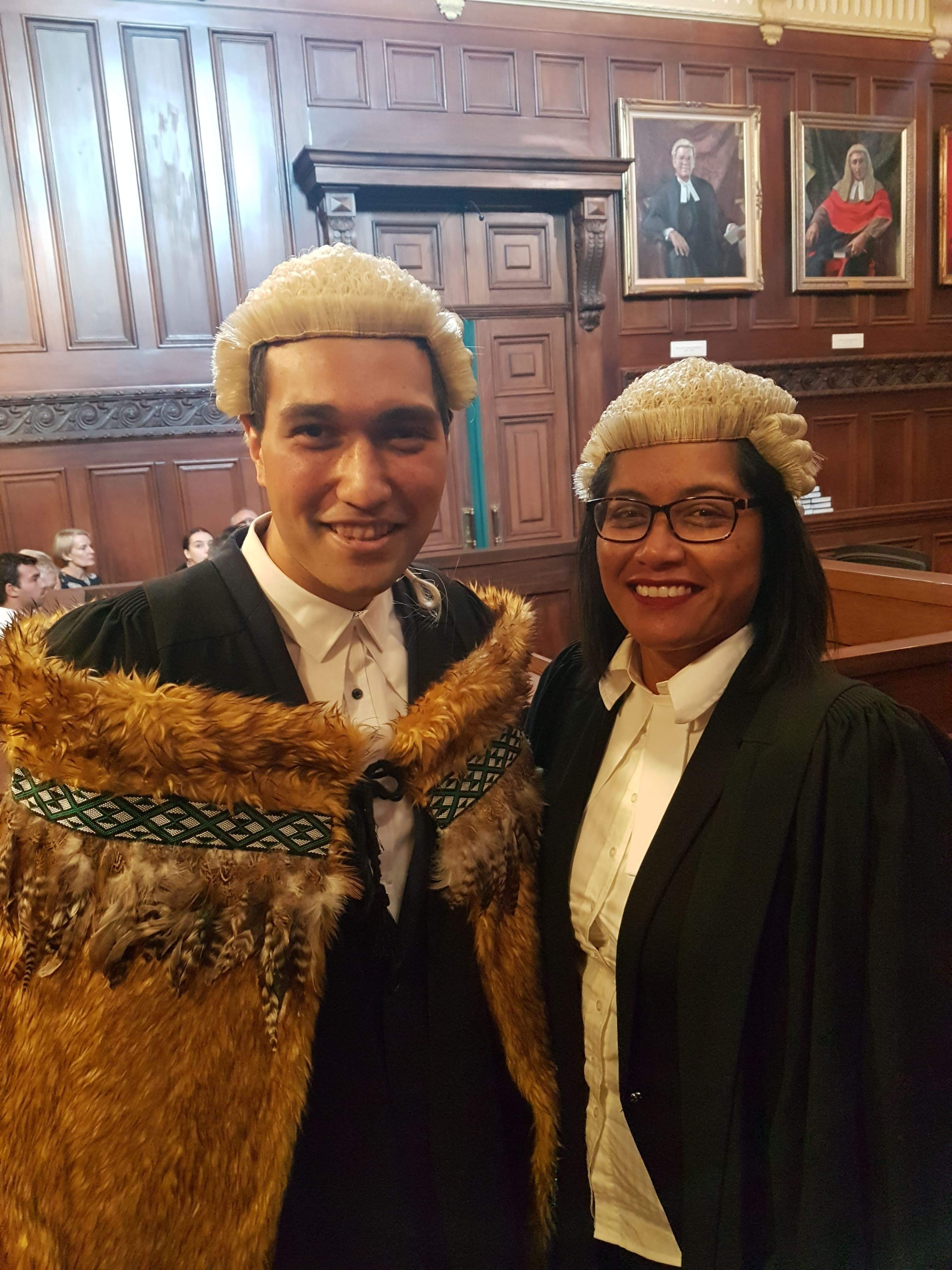 Duncan stands in a wood paneled courtroom next to a lady who is smiling. Both are wearing barrister wigs. Duncan is wearing a korowai (cloak)