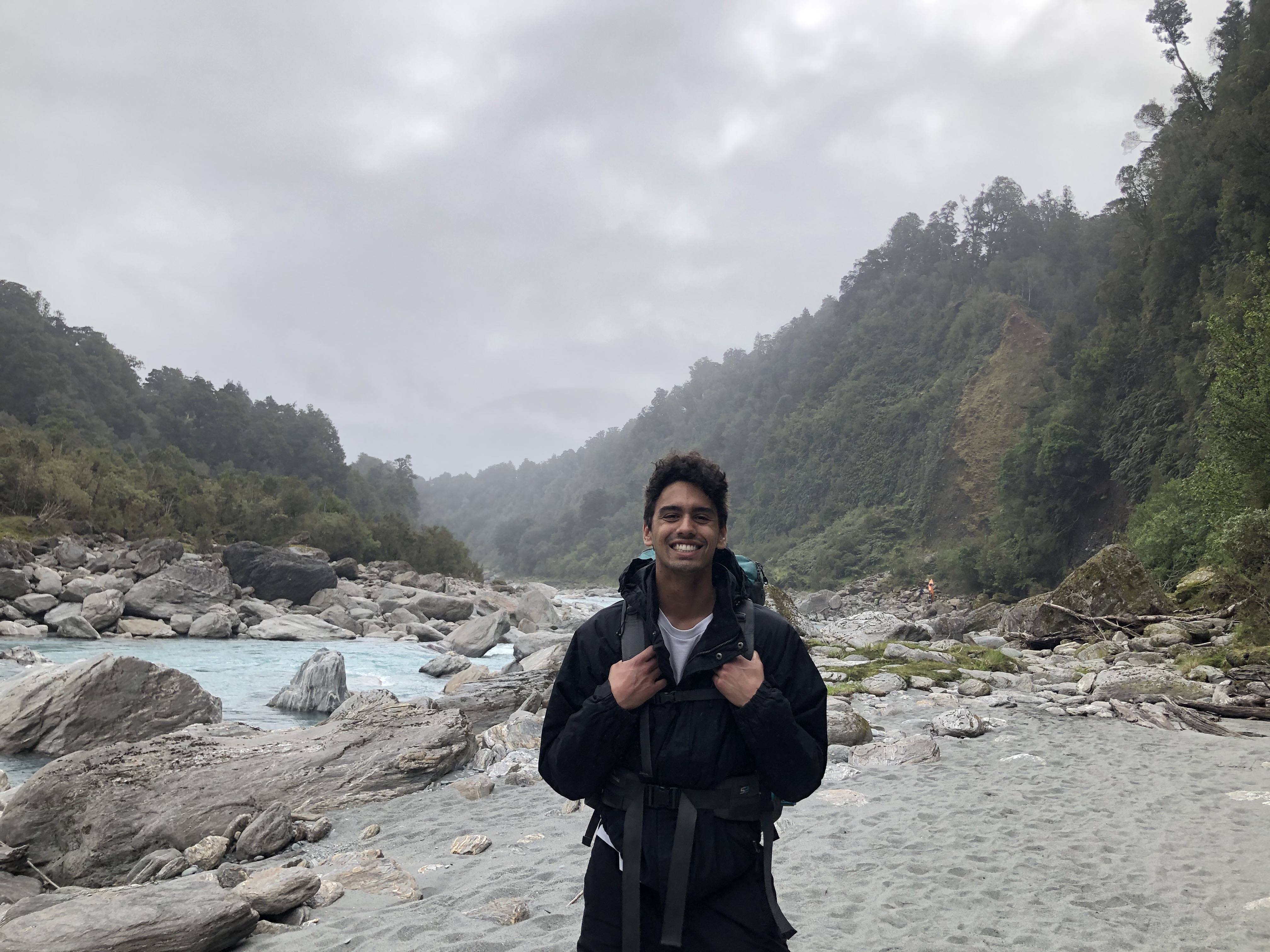 Young man standing and smiling at the camera whilst gripping the straps of his backpack which is on his shoulders. Behind him is a blue river, a rocky river bed and pine trees. There is a grey cloudy sky with mist starting to cover the trees.