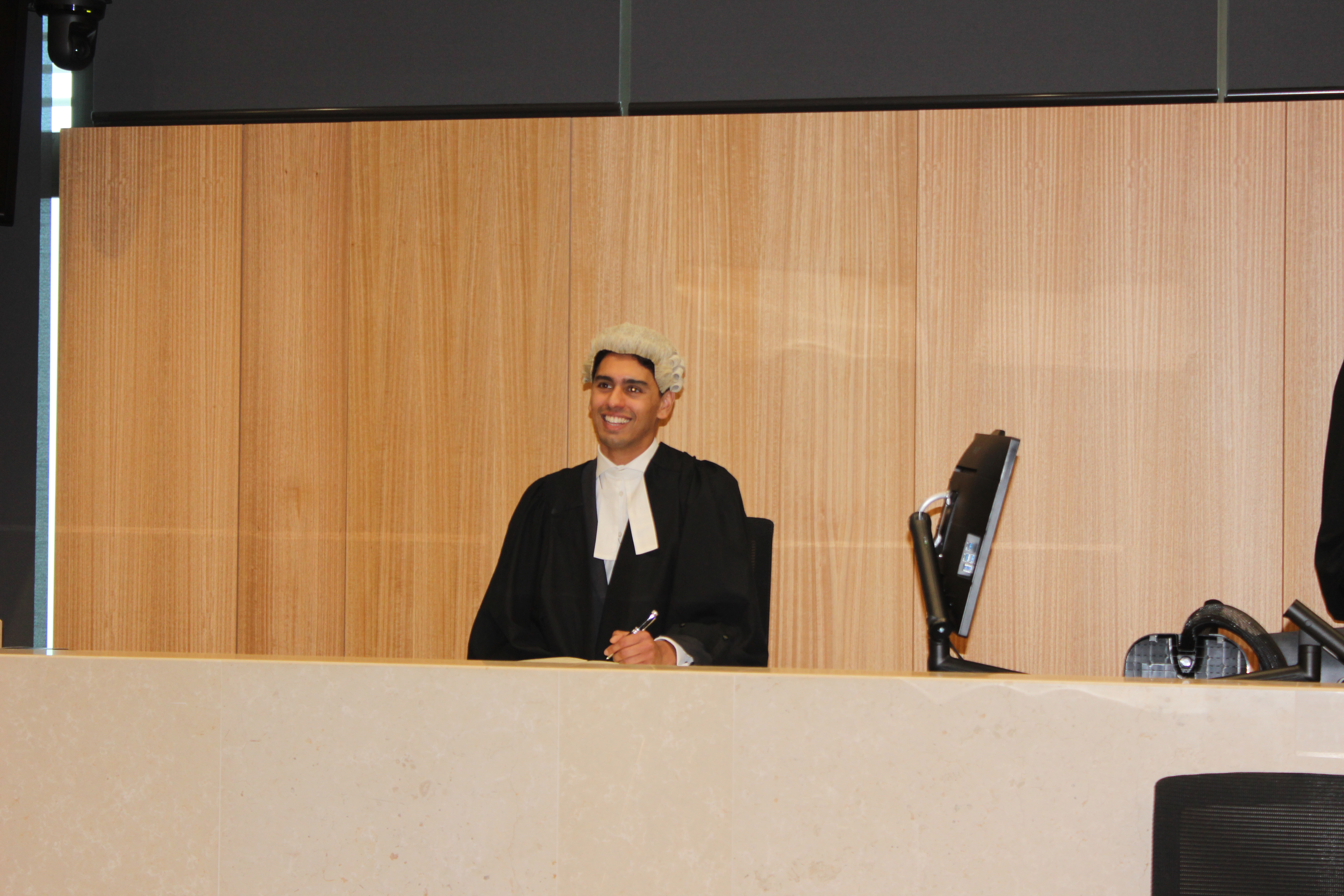 Ravi sits in the court room wearing a black gown and a while curly wig. He is smiling.