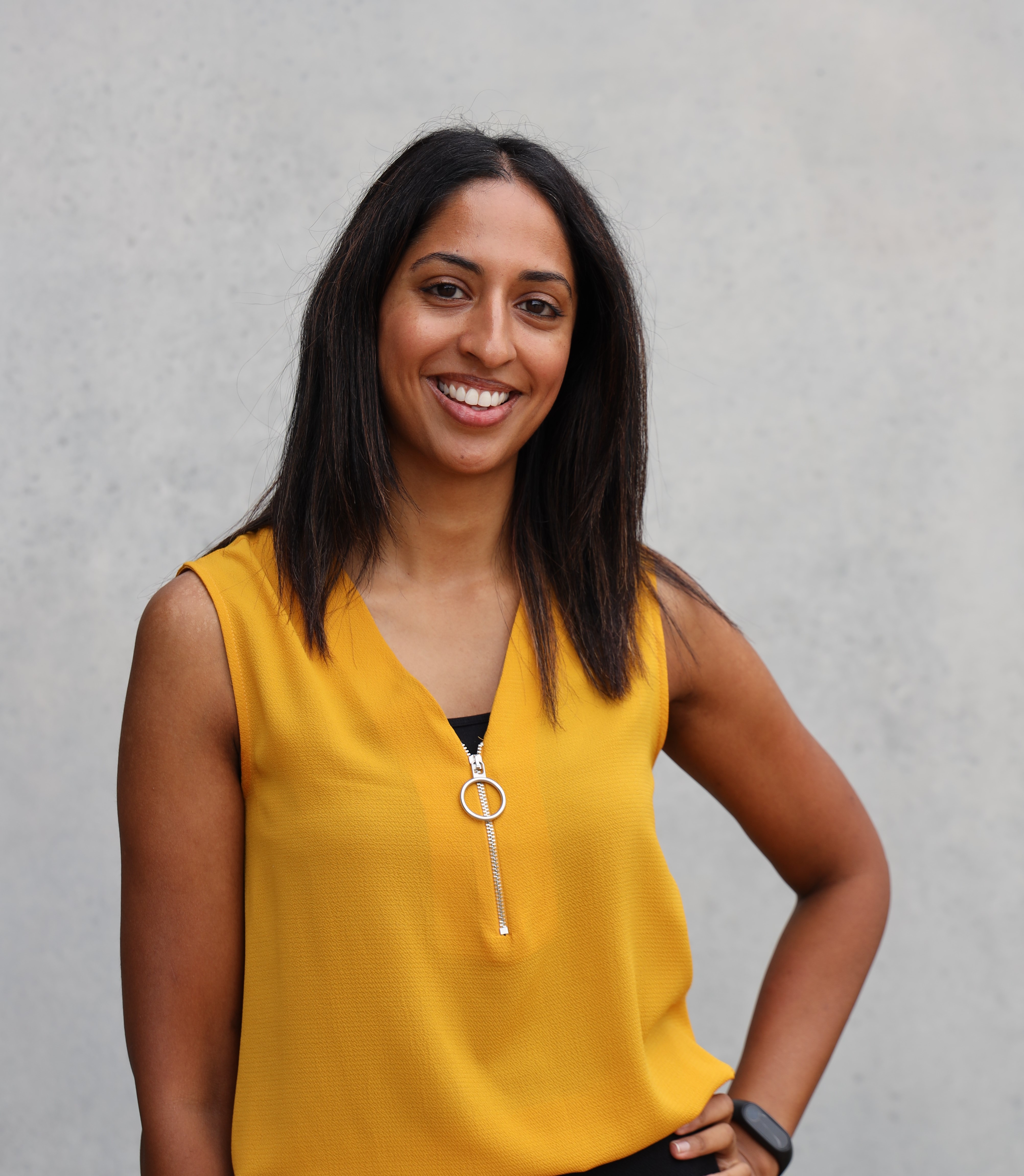 Farhanah Jeewa standing in front of a grey background wearing a bright yellow top, smiling and with her hand on her hip.